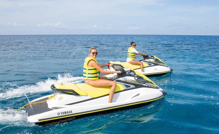 Two women riding jet skis in the ocean near a sailboat rental in Grand Cayman.