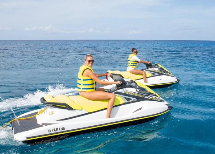Two women riding jet skis in the ocean near a sailboat rental in Grand Cayman.