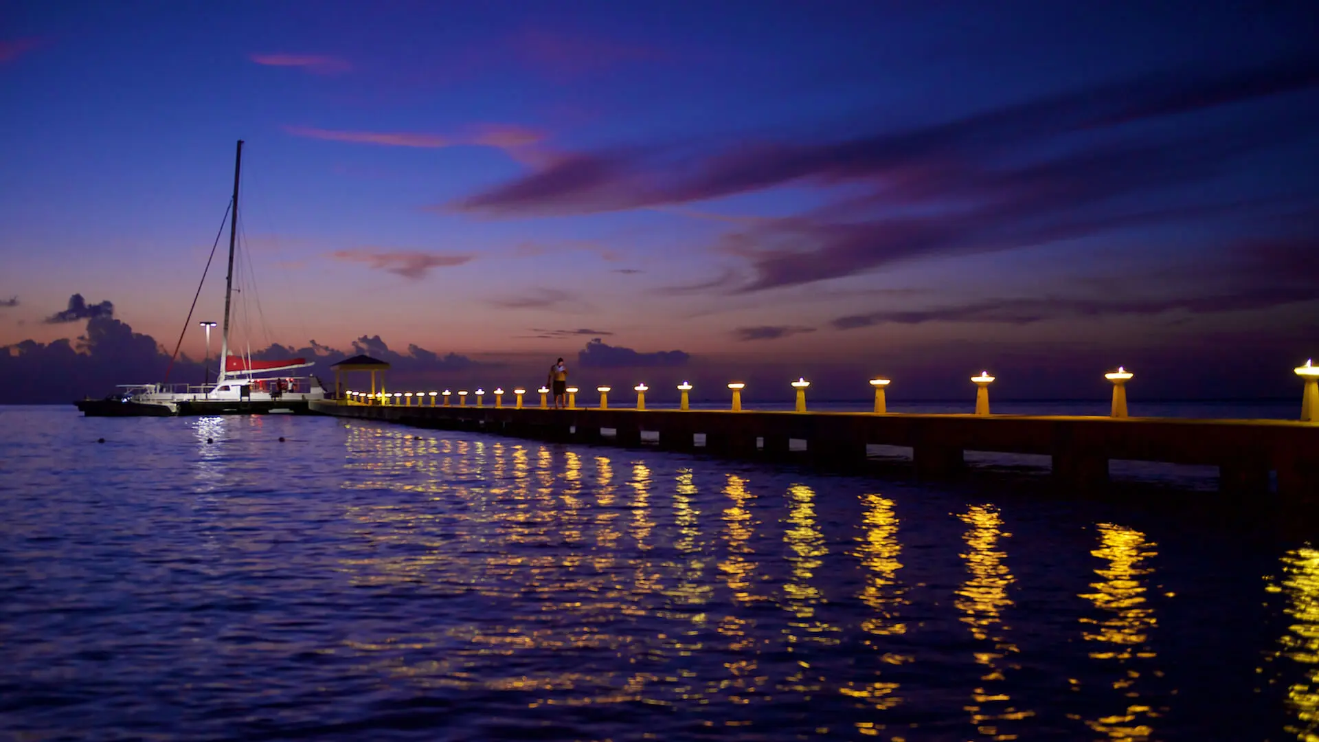 A sailboat docked at a pier during sunset, with lights reflecting on the calm water and a colorful sky in the background.