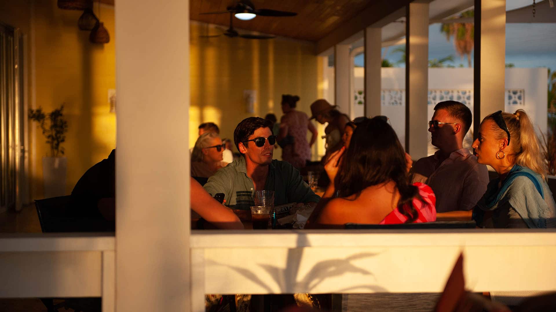 A group of people sit together at an outdoor cafe, conversing as sunlight casts shadows on the patio.