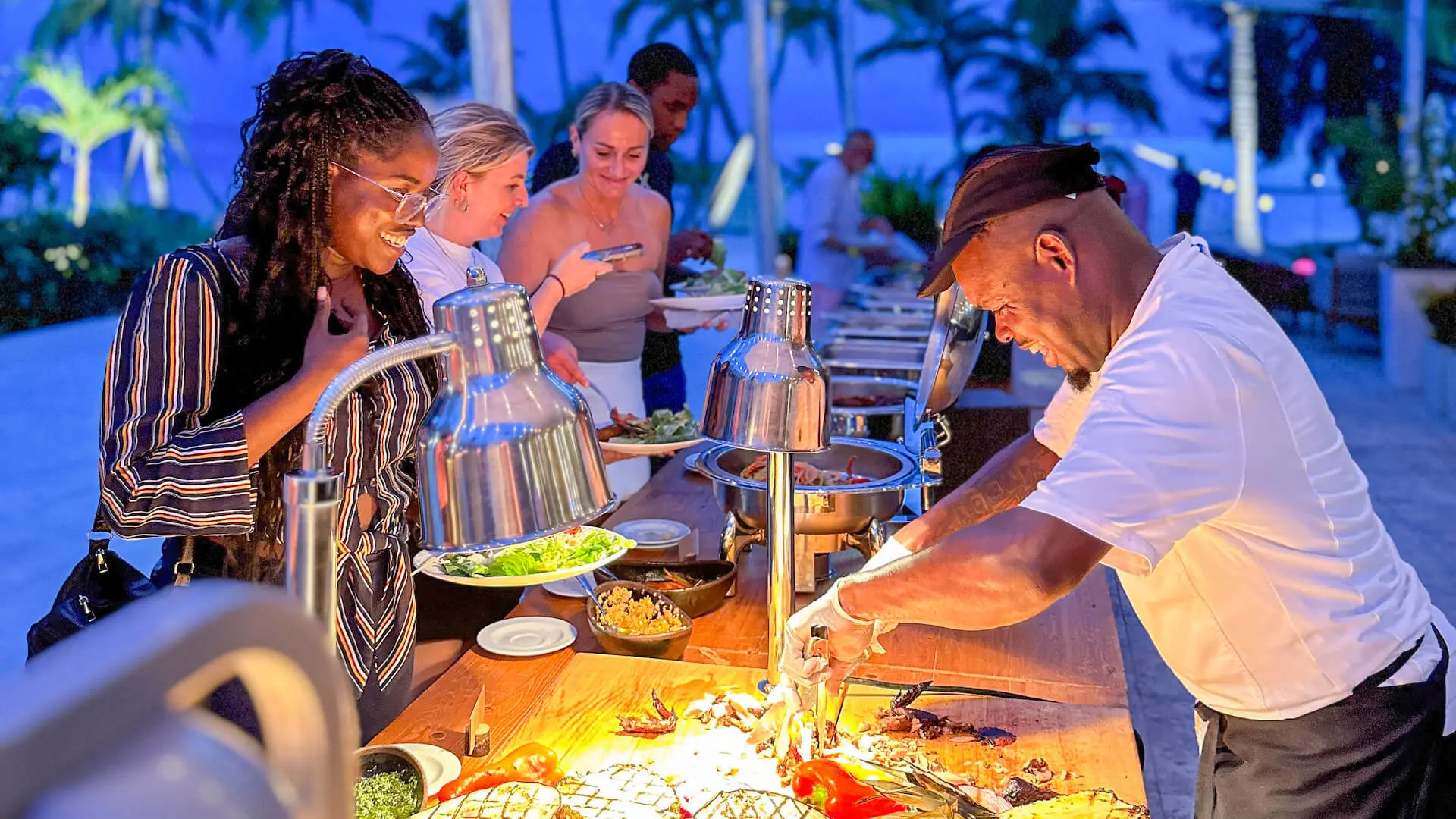 A chef serves food at an outdoor buffet to a group of people on a tropical evening.