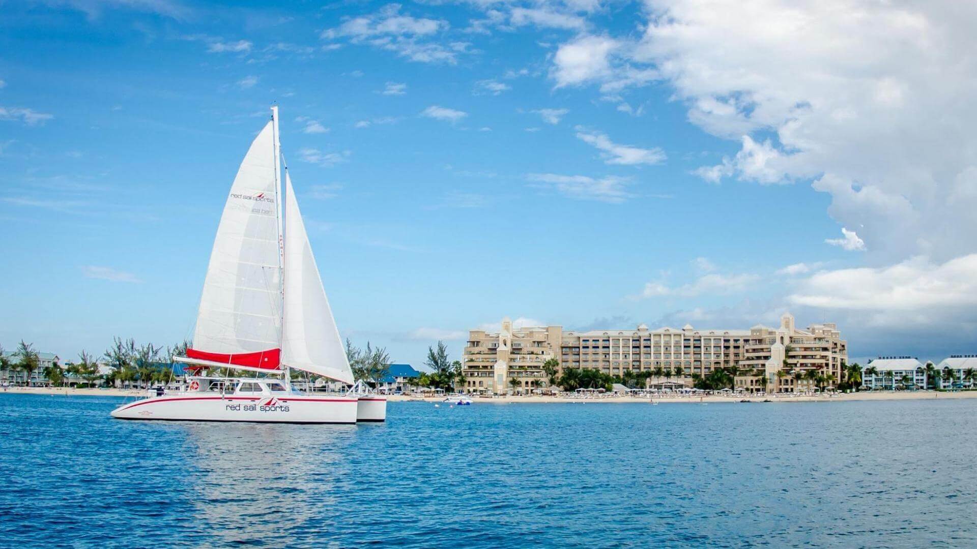 A white catamaran sails on blue water with a beach and resort buildings in the background under a partly cloudy sky.
