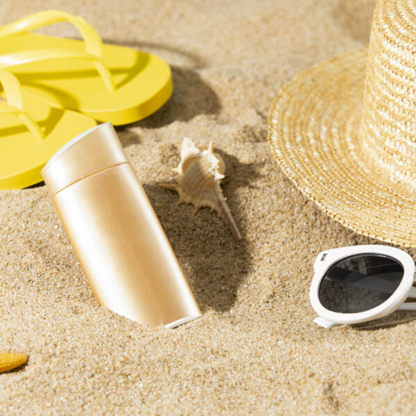 Beach essentials on sand: yellow flip-flops, sunscreen bottle, seashells, starfish, straw hat, and white sunglasses.