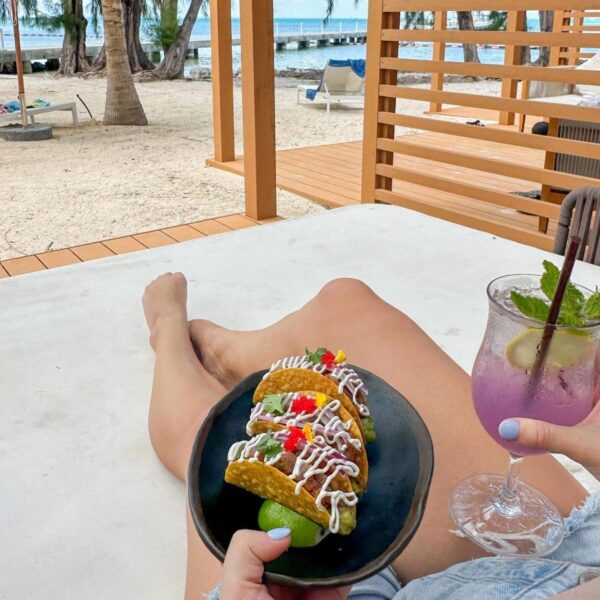 Relaxing at Rum Point, this person lounges on a beach chair, savoring tacos and a purple drink with lime and mint. With the sandy beach, swaying palm trees, and ocean in the background, it's one of the many fun things to do at this tropical paradise.