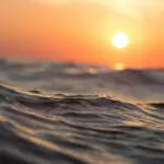 Sunset over the ocean with calm waves and an orange sky.