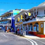 Street scene with colorful buildings and shops. A yellow building with a gallery is on the corner. People walk on the sidewalk, and cars drive on a curved road under a blue sky.