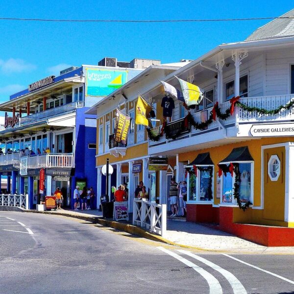 Street scene with colorful buildings and shops. A yellow building with a gallery is on the corner. People walk on the sidewalk, and cars drive on a curved road under a blue sky.