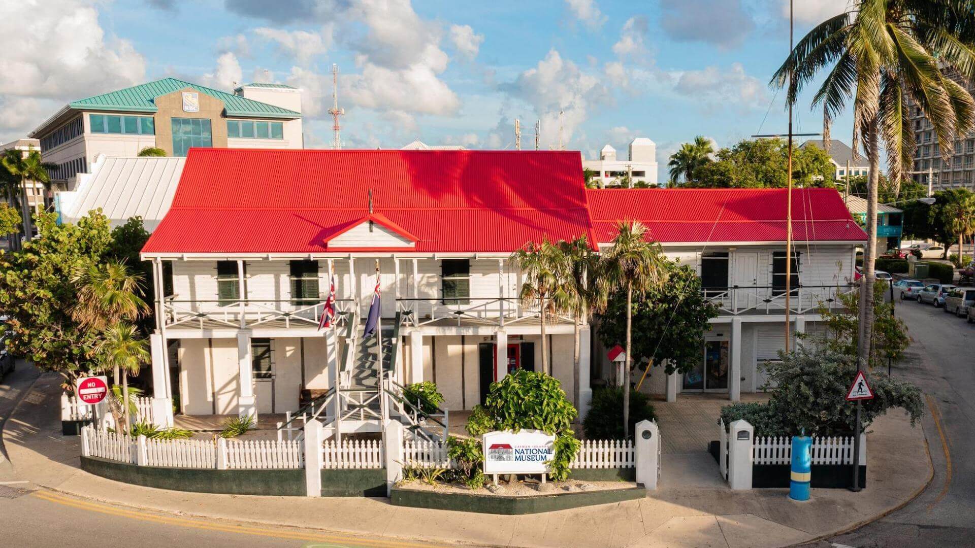 White colonial-style building with a red roof, surrounded by palm trees and a white picket fence. Flags are at the entrance. A road curves nearby under a partly cloudy sky.