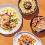 Four dishes on a wooden table: shrimp and avocado salad, baked eggplant, shrimp in sauce with bread slices, and grilled octopus with sauce.