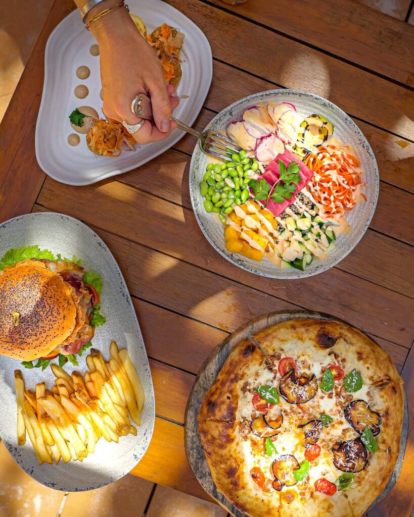 A table at Rum Deck with assorted dishes: a colorful poke bowl, a gourmet burger with fries, a pizza with toppings. A hand holding a fork reaches into the poke bowl.