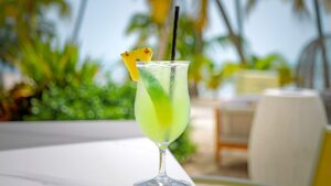 A green cocktail garnished with a pineapple slice and a straw sits on a white table outdoors, with tropical plants and palm trees blurred in the background.