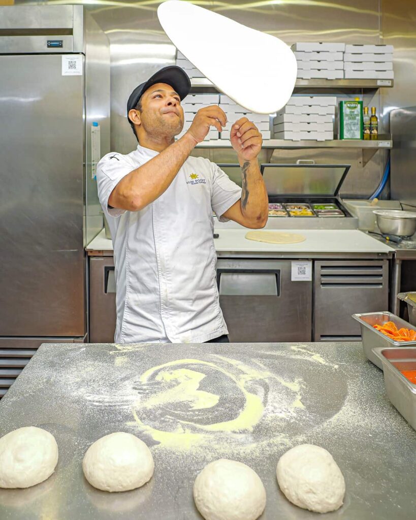 A chef in a white uniform tosses pizza dough in a commercial kitchen, with four dough balls and flour on a stainless steel counter in front of him.