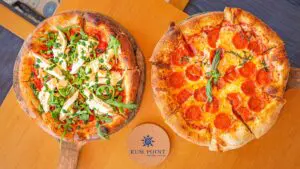 Two pizzas on wooden boards: one topped with arugula, cheese, and tomatoes, the other with pepperoni and basil, placed on a table with a Rum Point coaster in between.