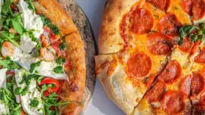 Two pizzas side by side: one topped with pepperoni slices and fresh basil, the other with arugula, burrata, shrimp, cherry tomatoes, and green pesto.